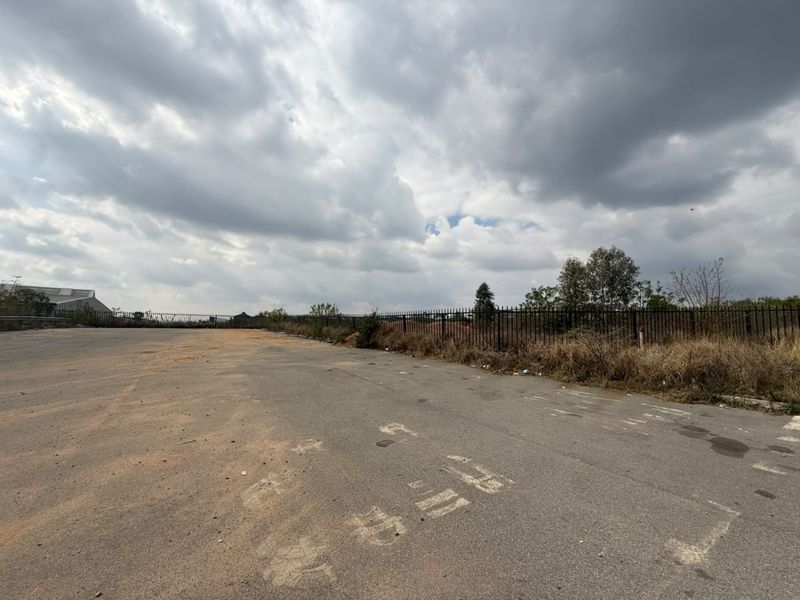 A vacant residential land plot featuring an asphalt surface and metal perimeter fencing under a cloudy sky.