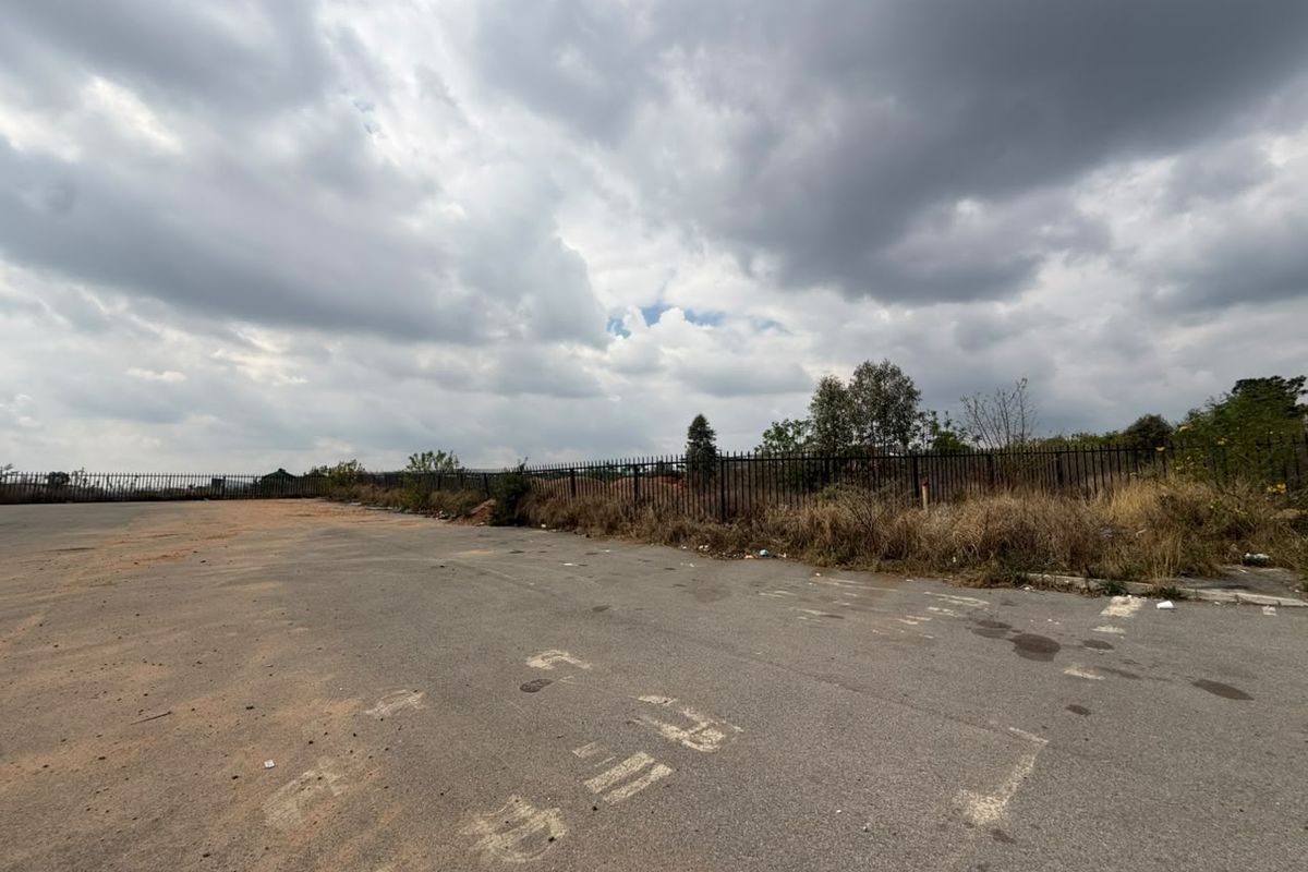 A vacant residential land plot featuring paved asphalt surfacing and perimeter metal fencing under an overcast sky.