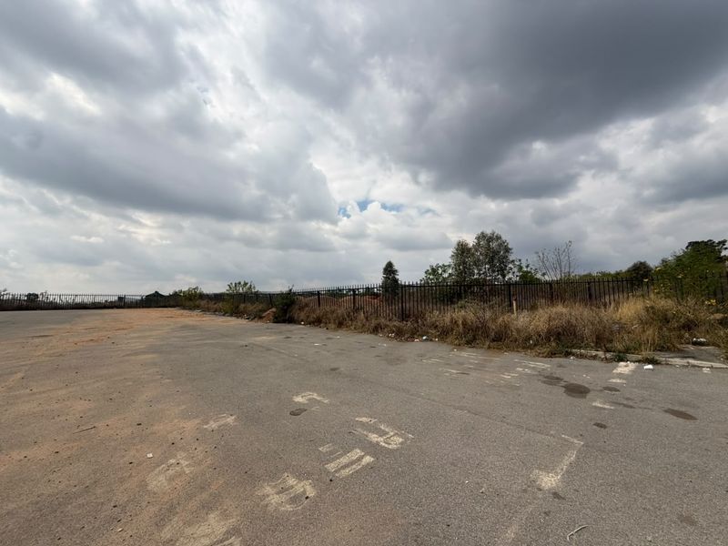 A vacant residential land plot featuring paved asphalt surfacing and perimeter metal fencing under an overcast sky.
