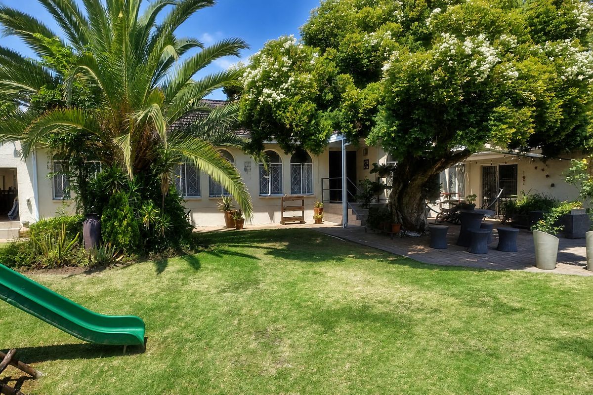 A spacious rear garden featuring a paved patio area, mature trees, and a well-maintained lawn, providing a pleasant outdoor living space.
