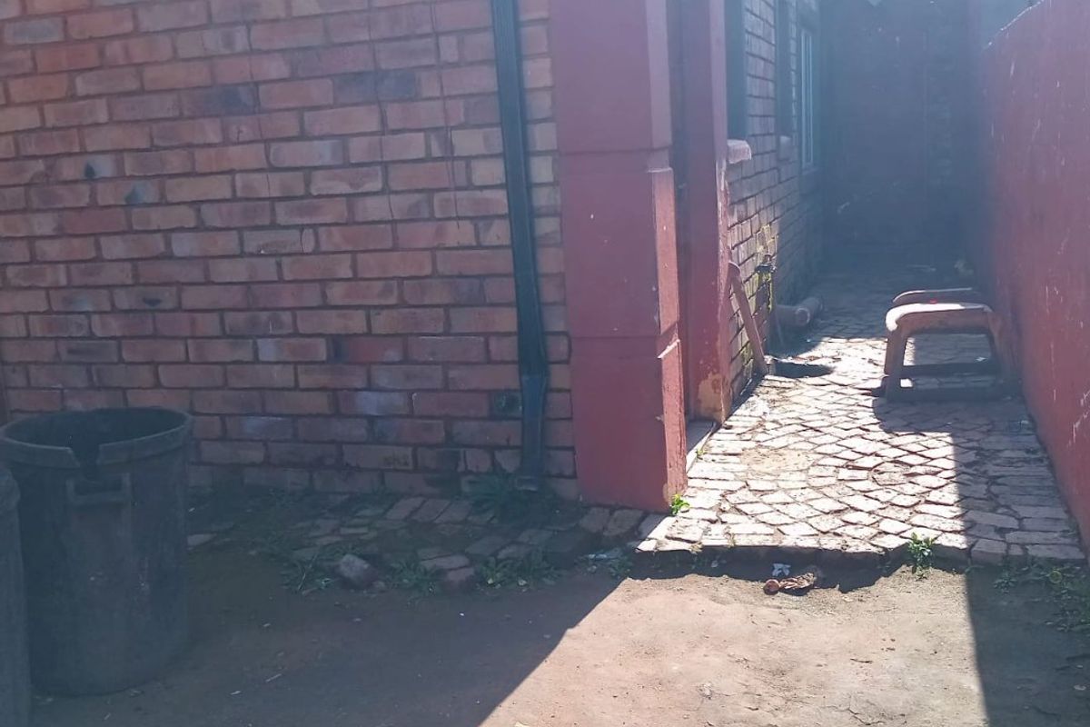A side exterior view of the property featuring a brick facade, paved walkway, and basic drainage infrastructure.