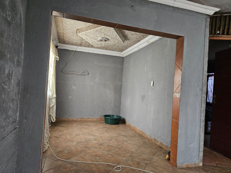 An empty living area featuring tiled flooring, decorative ceiling tiles, and coved cornicing, requiring cosmetic updates to the wall finishes and electrical fixtures.