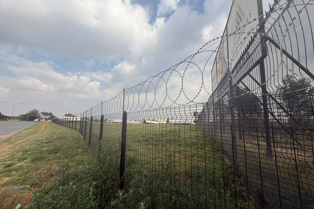 A vacant residential land plot secured with high-security mesh fencing, razor wire, and featuring a large advertising billboard along the perimeter.