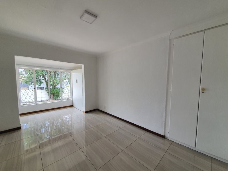 A bright, empty bedroom featuring neutral tiled flooring, built-in cupboards, and large windows with security bars, offering a clean and versatile living space.