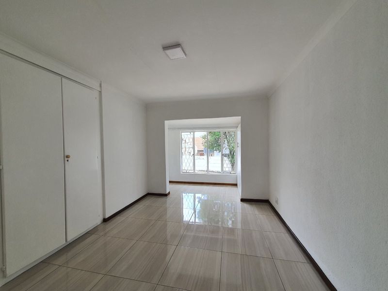 A bright, empty bedroom featuring neutral tiled flooring, built-in cupboards, and a window with security bars.