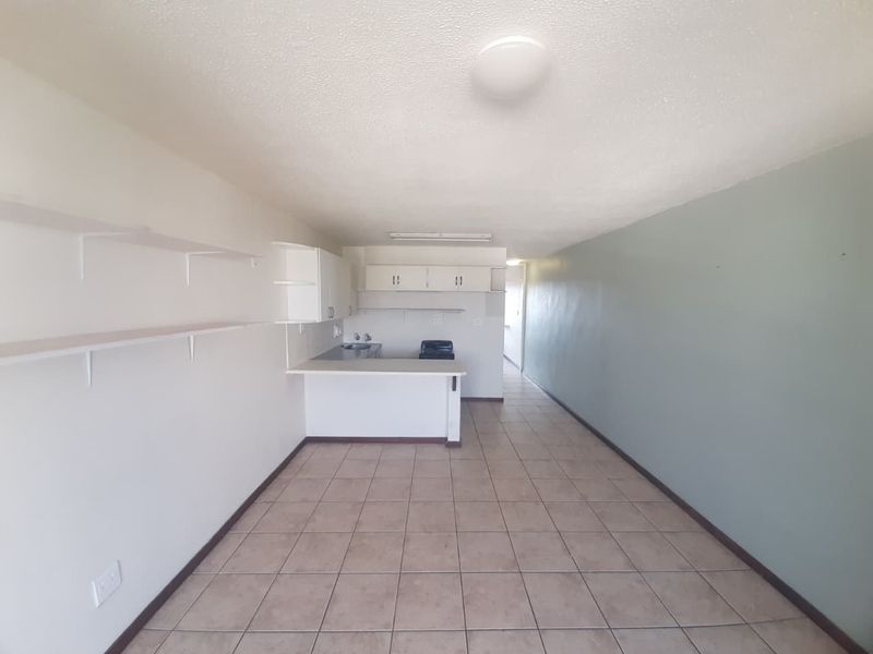 An open-plan living and kitchen area featuring tiled flooring, built-in cabinetry, a breakfast counter, and wall-mounted shelving in a neutral, vacant space.