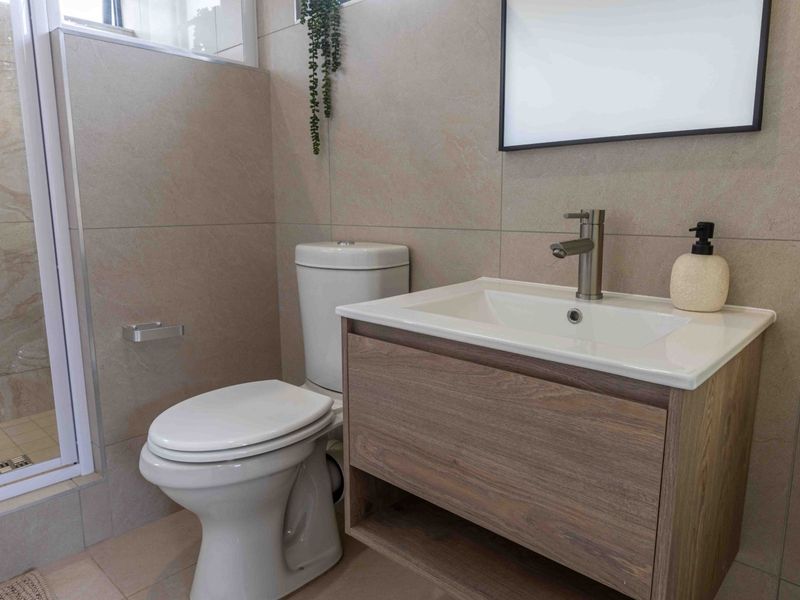 A modern bathroom featuring a wood-finish vanity cabinet, ceramic basin with mixer tap, toilet, and a glass-enclosed shower with neutral wall tiling.