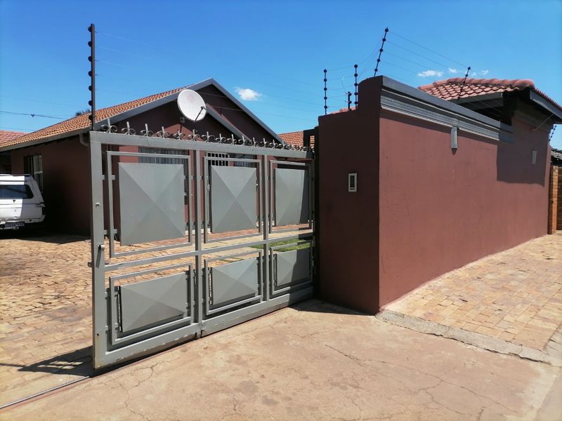 Front exterior view of the property featuring a paved driveway, secure metal gate, and perimeter wall with electric fencing.
