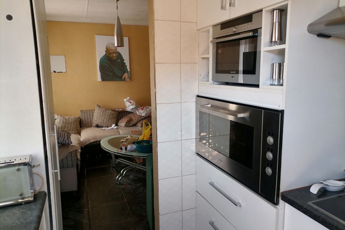 Compact kitchen featuring built-in oven and microwave, tiled splashback, and white cabinetry, opening into an adjacent living area.