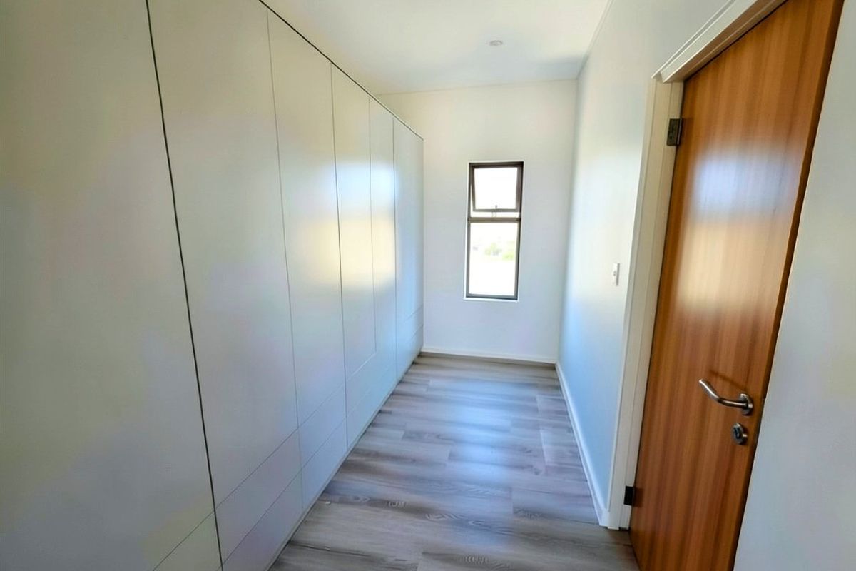 A clean, modern bedroom featuring extensive built-in floor-to-ceiling cupboards, light wood-look laminate flooring, and a single window providing natural light.