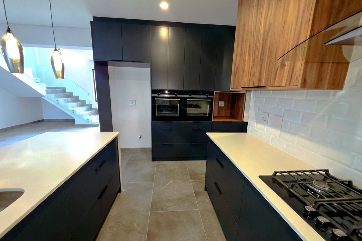 Modern kitchen featuring dark cabinetry, wood-tone accents, stone countertops, built-in ovens, and a gas hob, with an open-plan layout leading to a staircase.