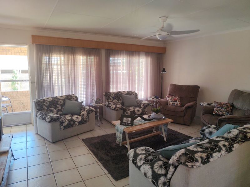 A comfortable living room featuring tiled flooring, a ceiling fan, and large windows with curtains, providing direct access to an outdoor patio area.