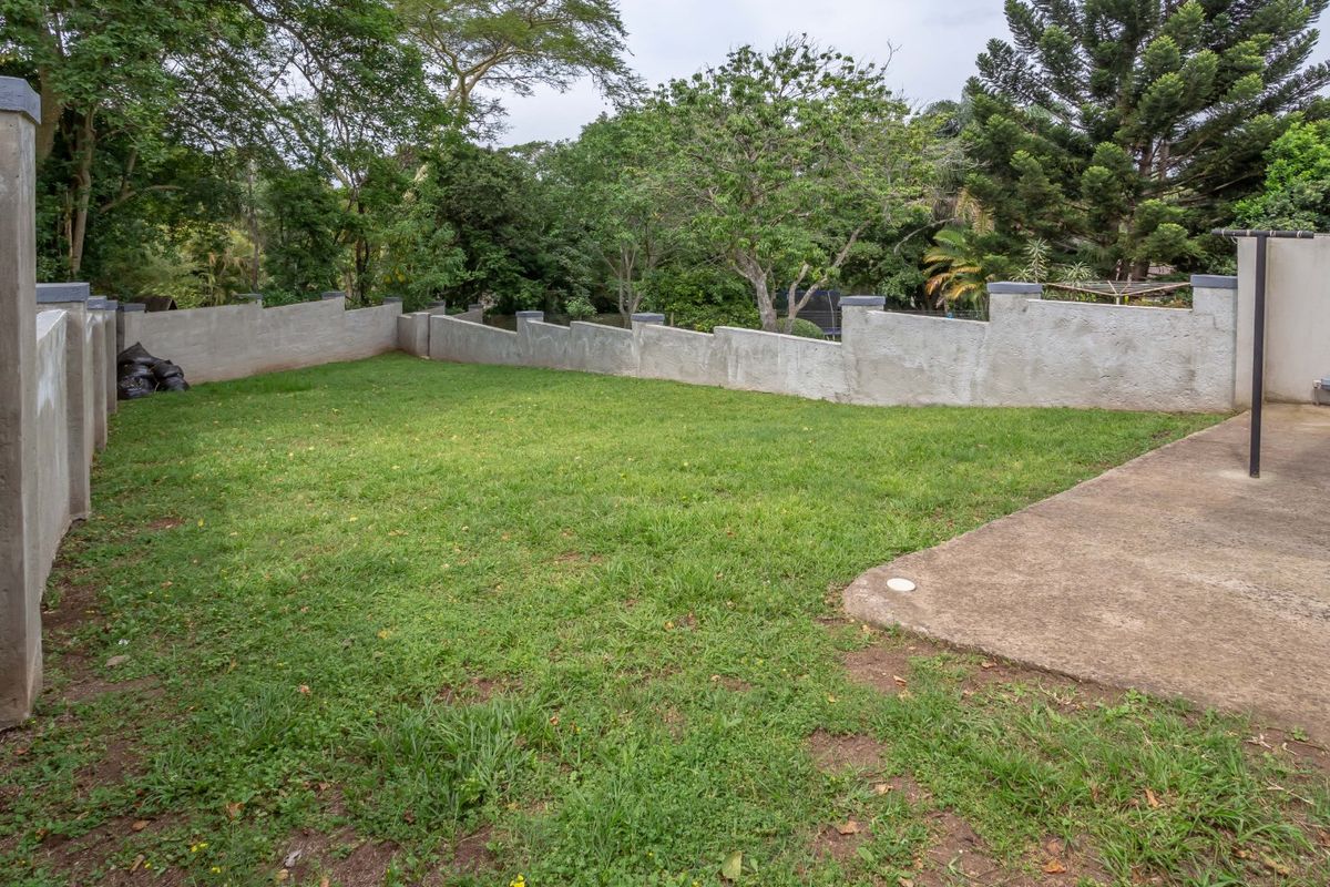A private rear garden featuring a lawn area, concrete patio, and perimeter boundary walls surrounded by mature trees.