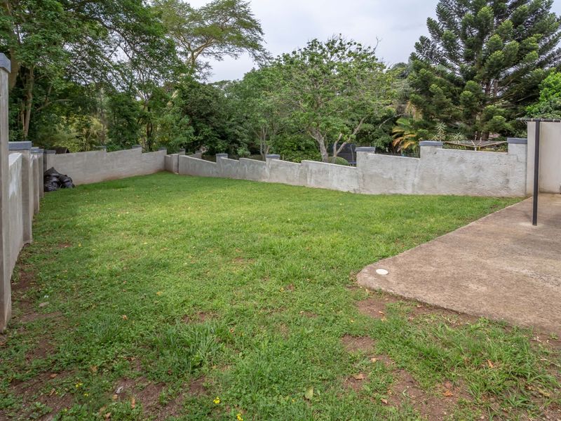 A private rear garden featuring a lawn area, concrete patio, and perimeter boundary walls surrounded by mature trees.