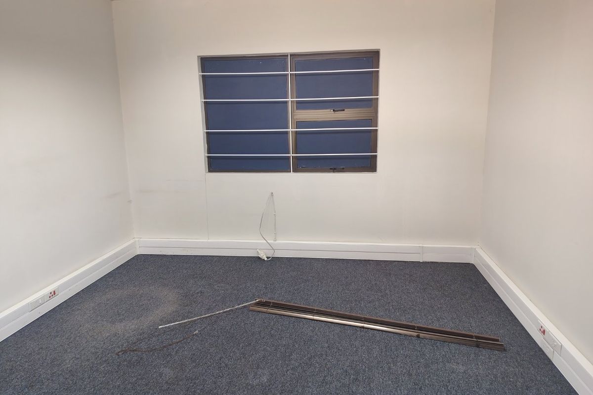 An empty office space featuring carpeted flooring, a window with security bars, and a suspended ceiling, currently requiring minor repairs and maintenance.