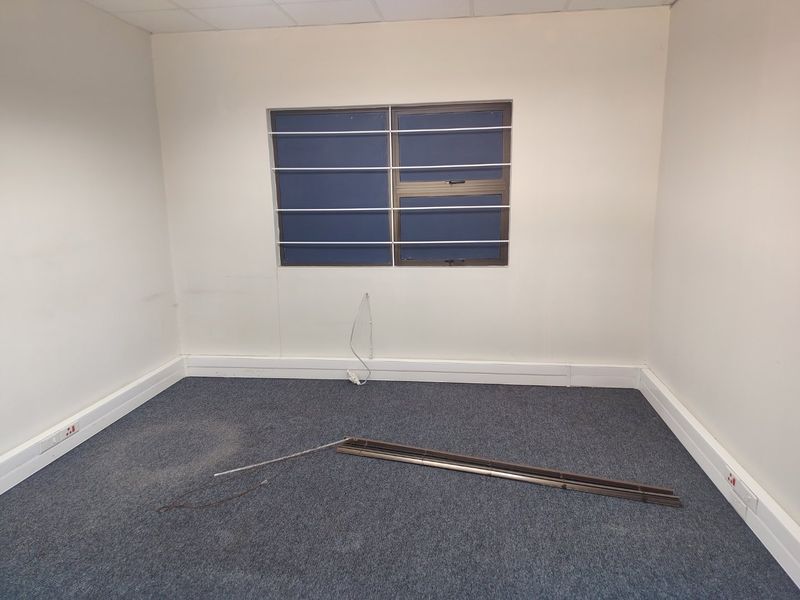 An empty office space featuring carpeted flooring, a window with security bars, and a suspended ceiling, currently requiring minor repairs and maintenance.