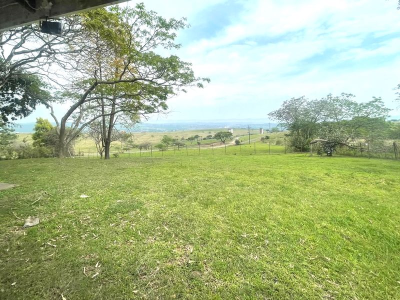 Expansive rural landscape view featuring open green fields, mature trees, and perimeter wire fencing, captured from an elevated vantage point.
