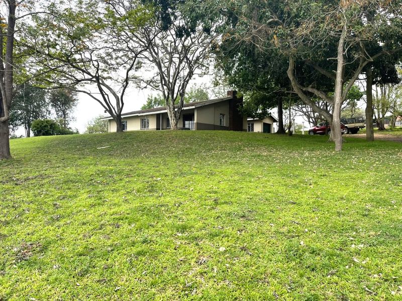 A view of the rear elevation of the house, featuring a sloped roof, brick chimney, and a spacious, tree-lined garden.