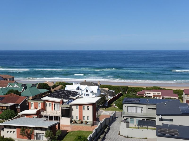 An elevated aerial view of a coastal residential neighborhood featuring homes with solar panels, overlooking the ocean and sandy beach.