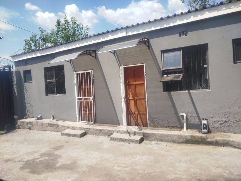Rear exterior view of a residential building featuring concrete apron, timber doors with security gates, and window awnings, showing signs of wear and maintenance needs.