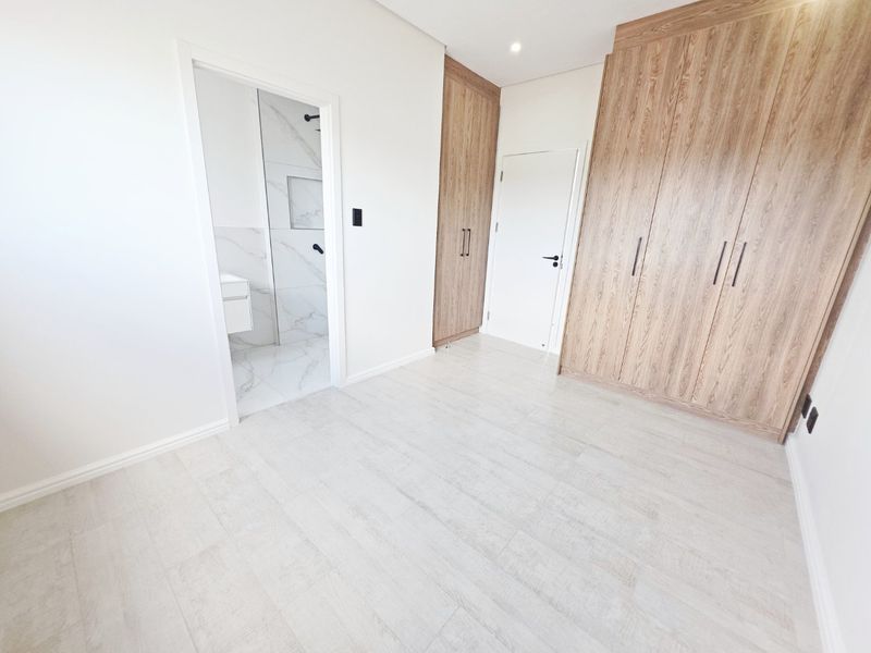 A modern, empty bedroom featuring light-toned tiled flooring, extensive built-in wooden cupboards, and direct access to an en-suite bathroom.