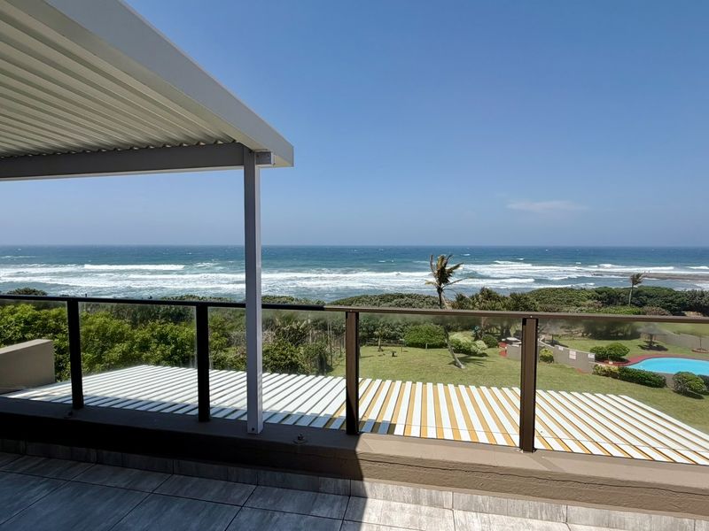 A spacious covered balcony featuring a modern glass balustrade and tiled flooring, offering expansive ocean and coastal views.