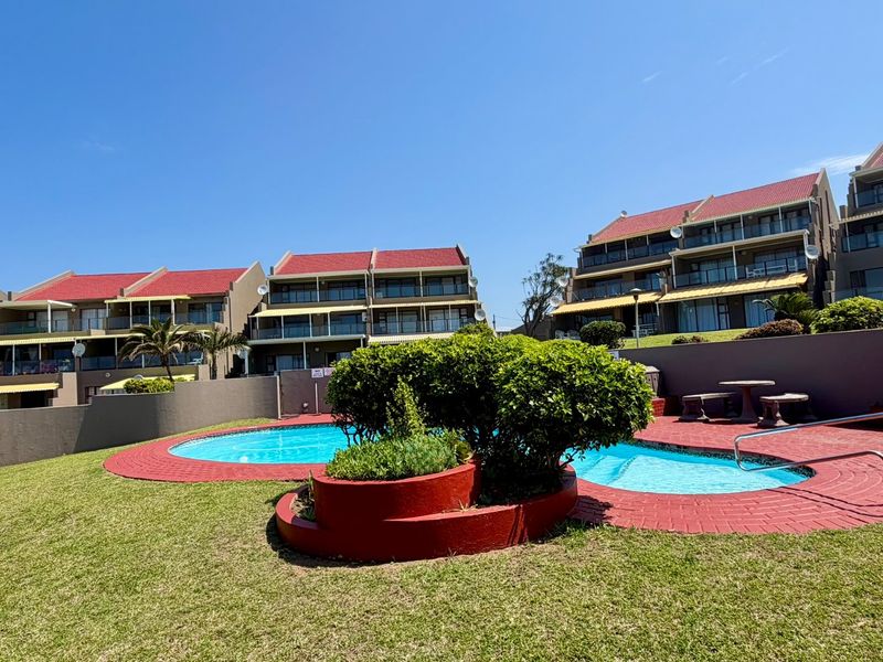 A well-maintained communal pool area featuring a swimming pool, brick paving, and landscaped garden beds, set against the backdrop of multi-story apartment buildings.
