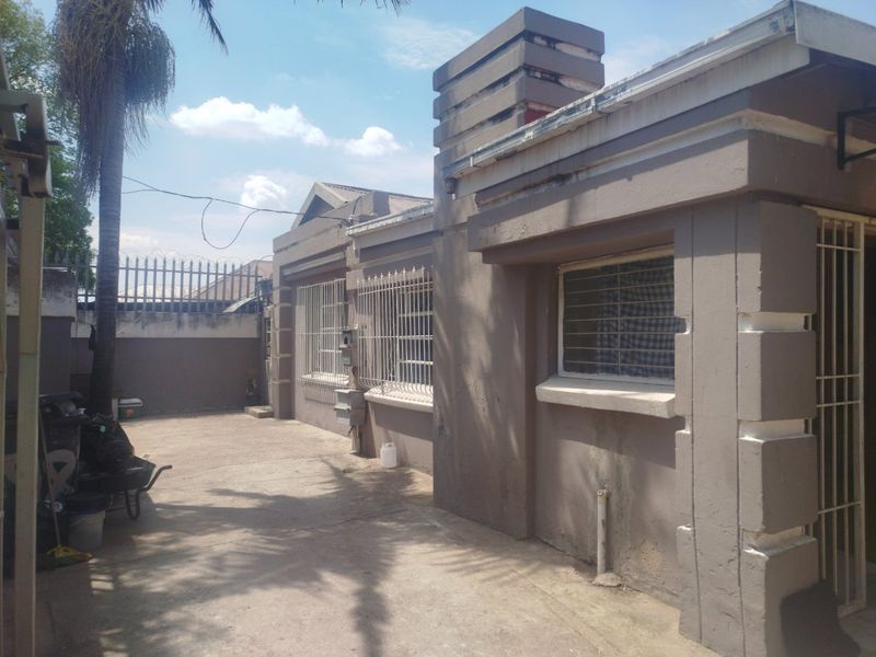 Rear exterior view of the property featuring concrete paving, security bars on windows, and a chimney stack, showing signs of wear on the exterior walls.