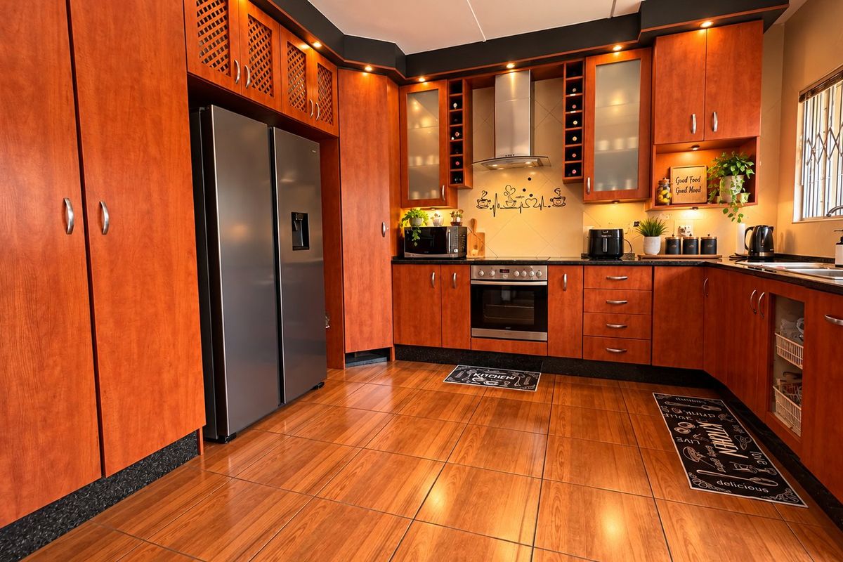 A well-maintained kitchen featuring warm-toned wooden cabinetry, integrated oven and extractor fan, dark countertops, and tiled flooring with a window security gate.