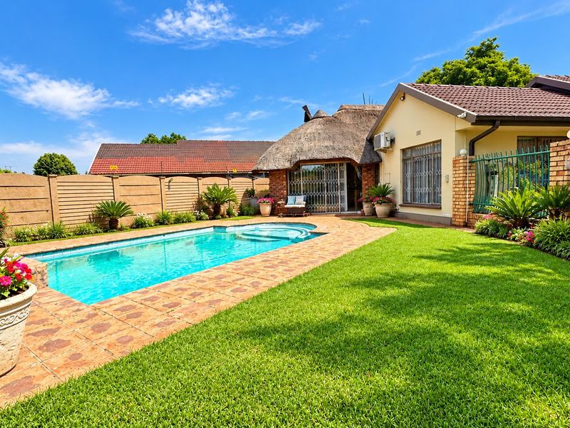 A well-maintained backyard featuring a swimming pool, paved surround, lush lawn, and a thatched-roof structure, enclosed by brick boundary walls.