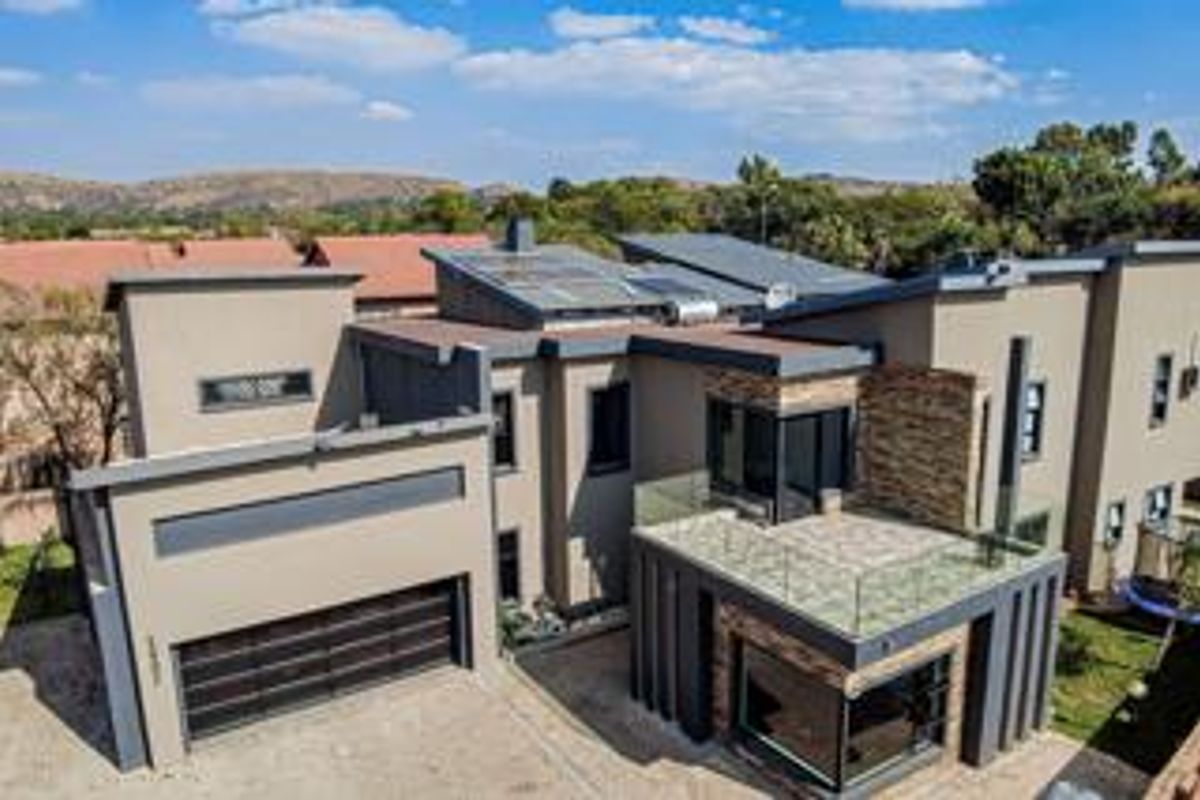 Aerial view of a modern multi-level residence featuring stone cladding, solar energy systems, a spacious paved driveway, and a rooftop terrace with glass balustrades.