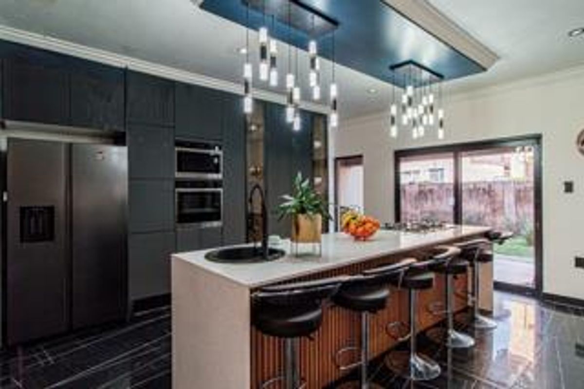 Modern kitchen featuring a large island with stone countertops, integrated appliances, dark cabinetry, and stylish pendant lighting, opening onto an outdoor area.