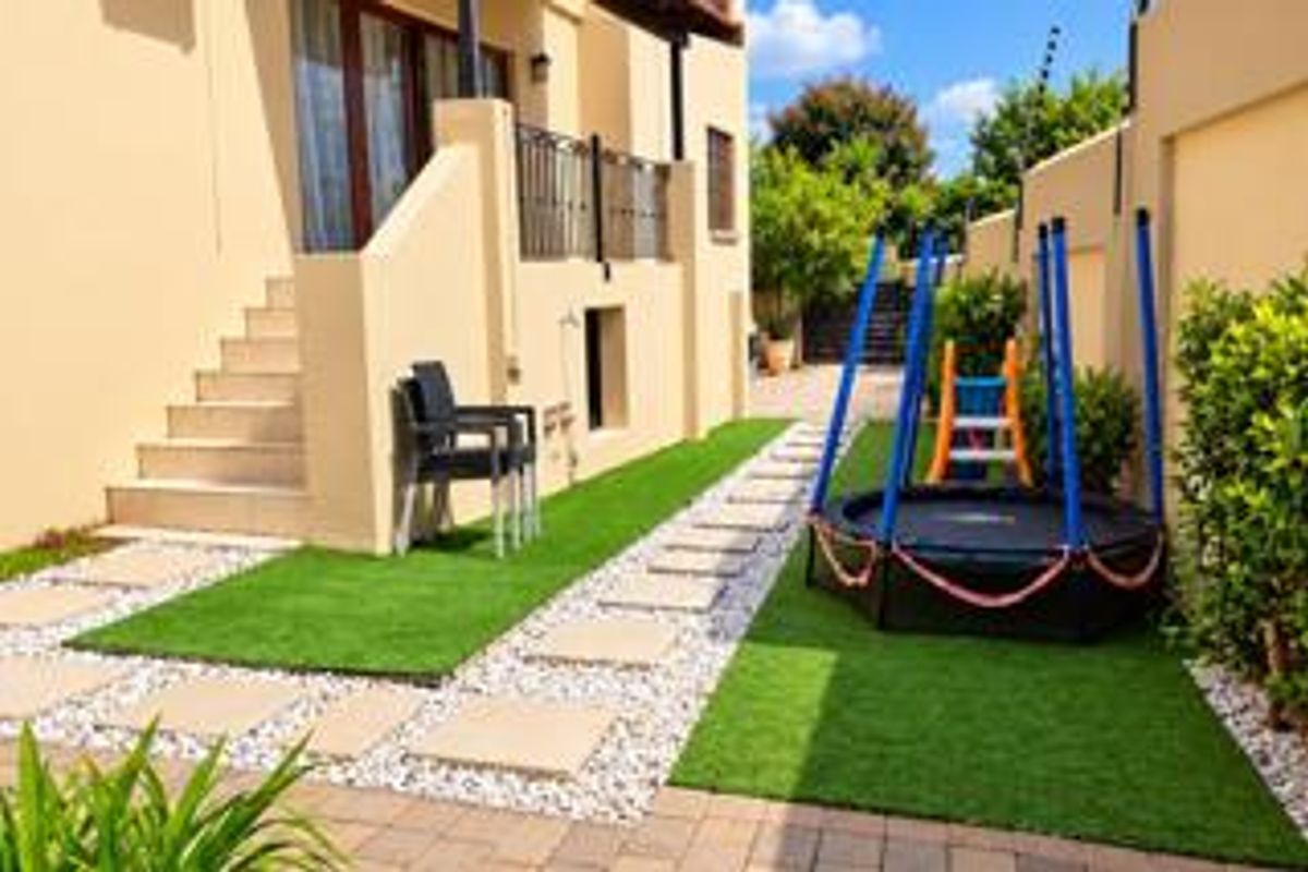 A well-maintained rear garden area featuring artificial lawn, stone paving pathways, and a concrete staircase leading to the upper level of the house.