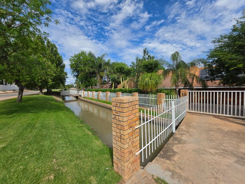 Front exterior view featuring a brick boundary wall, metal fencing, concrete driveway, and a water canal with a pedestrian bridge.