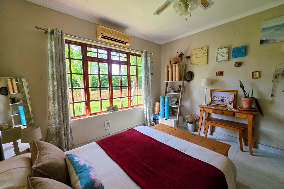A well-lit bedroom featuring tiled flooring, a ceiling fan, and a wall-mounted air conditioning unit, with large windows overlooking the garden.