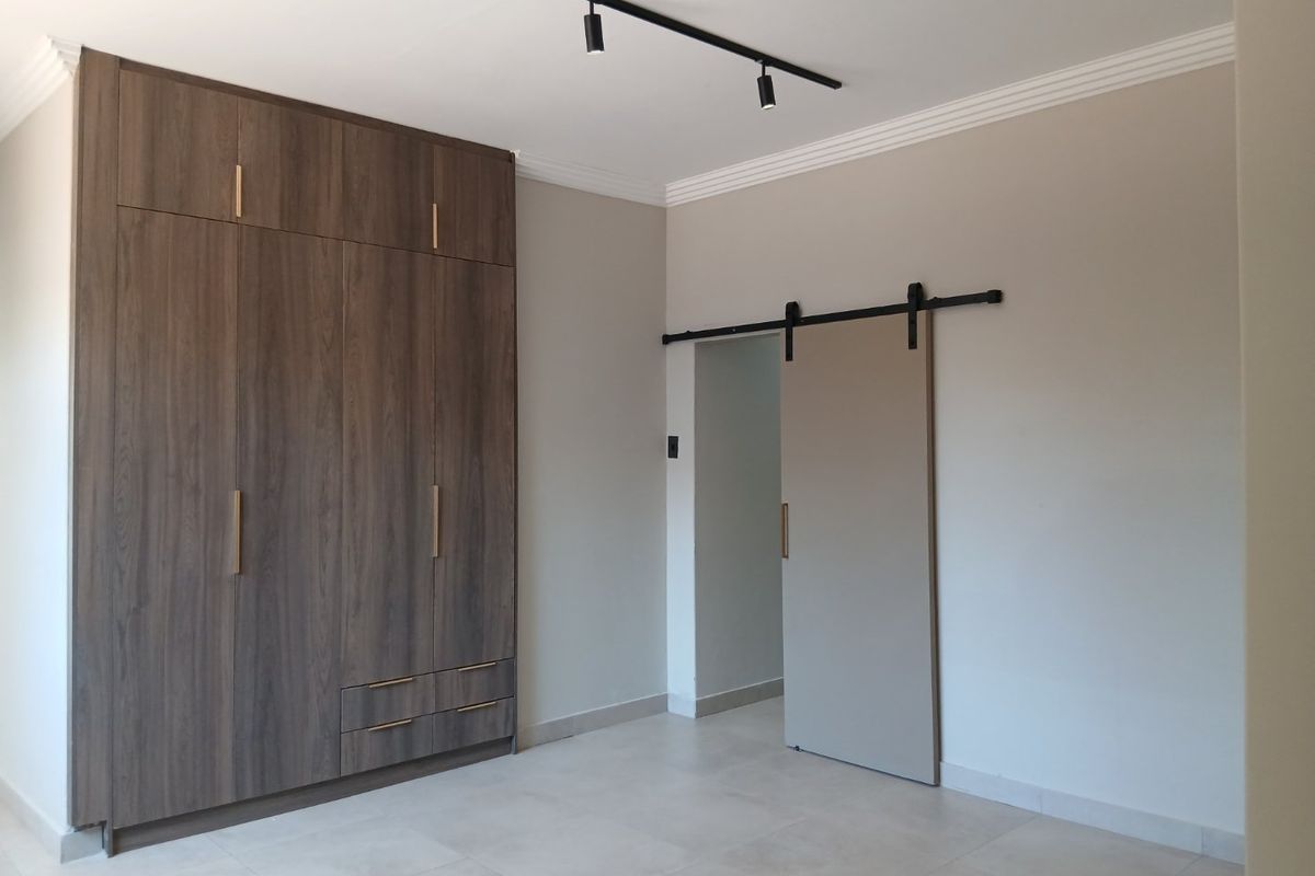 A clean, modern bedroom featuring floor-to-ceiling built-in cupboards, a contemporary sliding barn door, track lighting, and neutral tiled flooring.