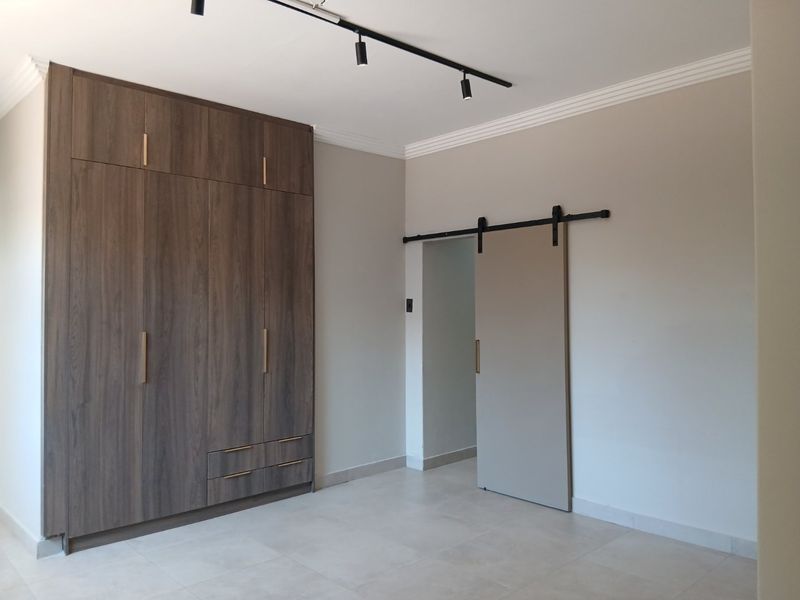 A clean, modern bedroom featuring floor-to-ceiling built-in cupboards, a contemporary sliding barn door, track lighting, and neutral tiled flooring.