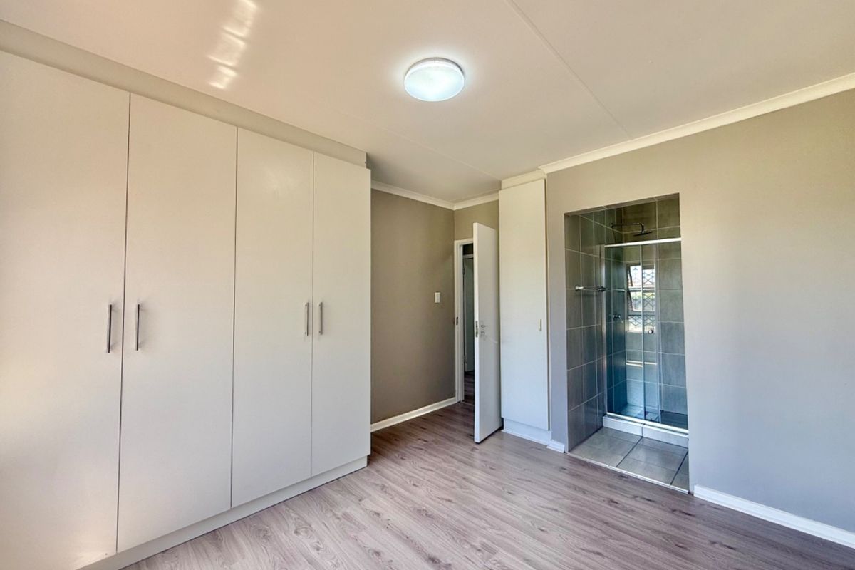 A clean, empty bedroom featuring modern laminate flooring, extensive built-in cupboards, and a convenient en-suite shower with tiled finishes.