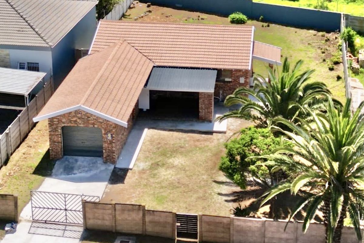 Aerial view of a single-story brick house featuring a tiled roof, attached garage, perimeter wall, and established garden with palm trees.