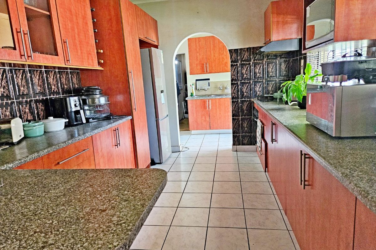 A functional kitchen featuring wood-finish cabinetry, granite countertops, and a tiled backsplash, with an adjoining scullery area accessible through an archway.