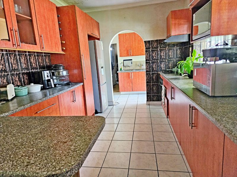 A functional kitchen featuring wood-finish cabinetry, granite countertops, and a tiled backsplash, with an adjoining scullery area accessible through an archway.