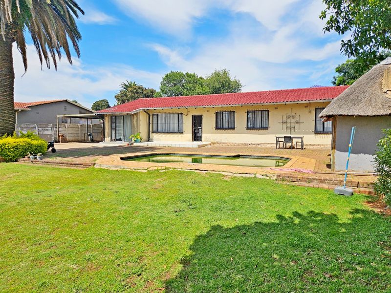 Rear view of the property featuring a swimming pool, paved patio area, and a small thatched lapa set within a spacious garden.