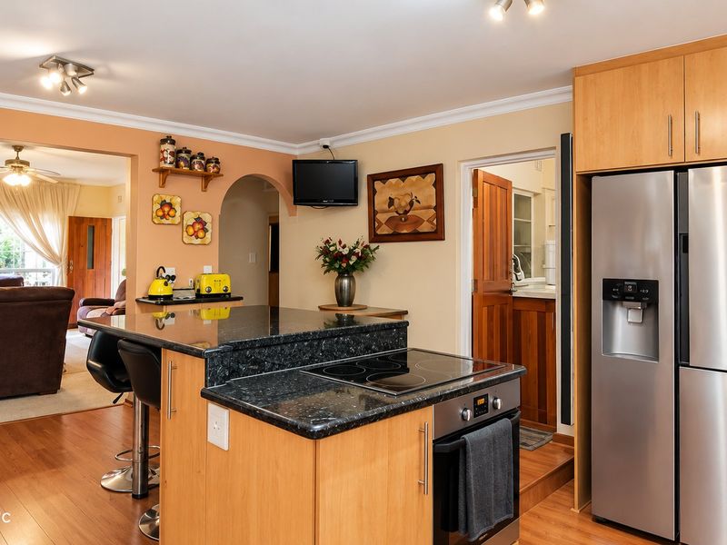Open plan kitchen featuring a central island with granite countertops, built-in hob and oven, and wooden cabinetry, flowing into the adjacent living area.