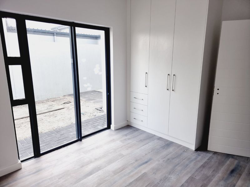 A bright, modern bedroom featuring built-in cupboards, light-toned laminate flooring, and a large sliding glass door providing direct access to the rear outdoor area.