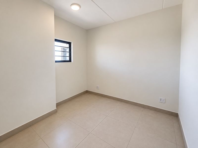 A clean, empty bedroom featuring neutral tiled flooring, a small window with security bars, and modern electrical finishes.