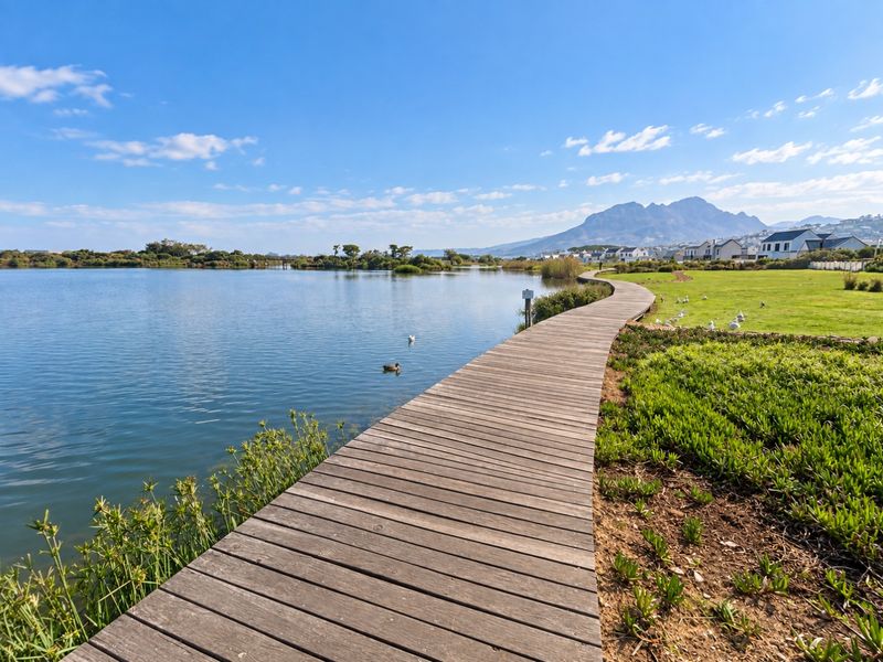 Scenic view of a wooden boardwalk winding along a tranquil lake, surrounded by landscaped lawns and mountain vistas.
