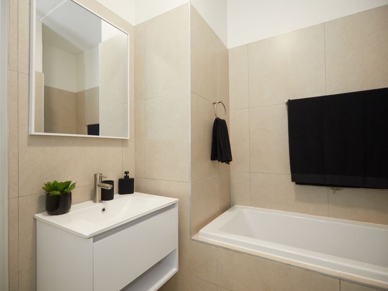 A modern bathroom featuring a white vanity cabinet with basin, a wall-mounted mirror, and a built-in bathtub with neutral ceramic wall tiling.