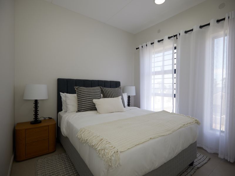 A well-maintained bedroom featuring neutral walls, tiled flooring, and a window fitted with security bars and sheer curtains.