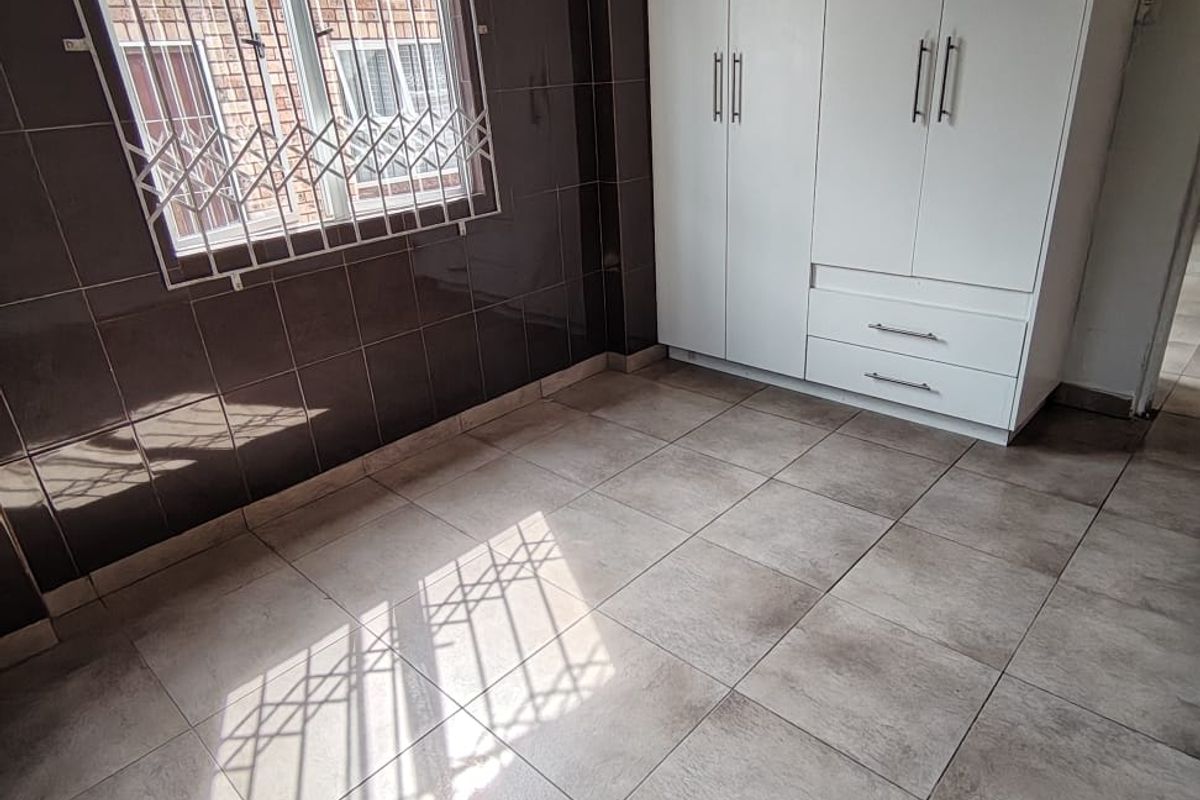 A clean, empty bedroom featuring tiled flooring, dark wall tiles, and a white built-in wardrobe with security bars on the window.