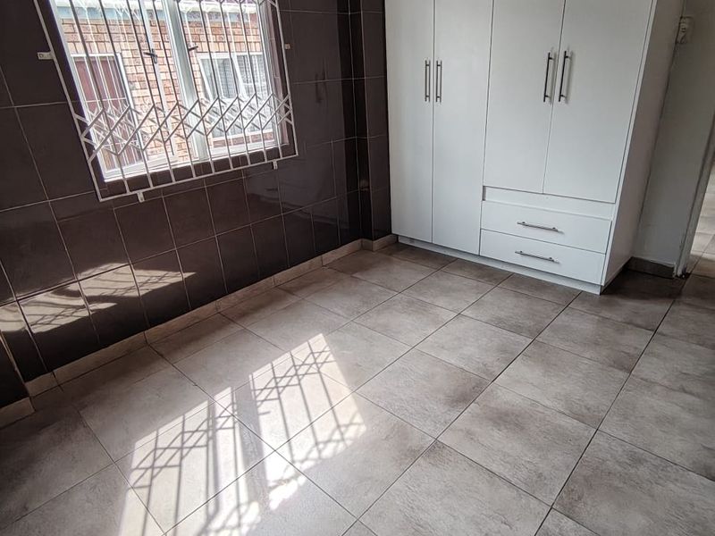 A clean, empty bedroom featuring tiled flooring, dark wall tiles, and a white built-in wardrobe with security bars on the window.